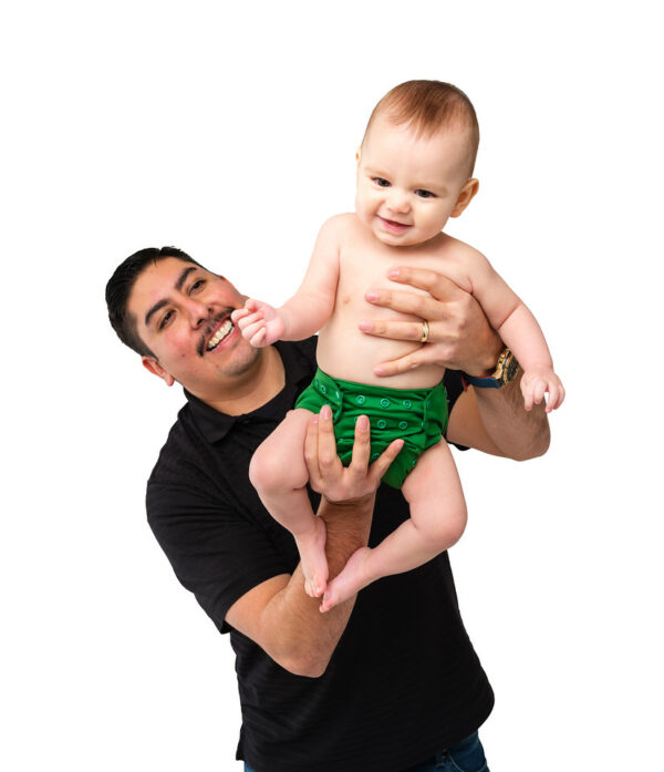 Dad with dark hair and mustache holding up a 6 month old baby wearing an all-in-one diaper in green. Baby and dad are smiling at each other